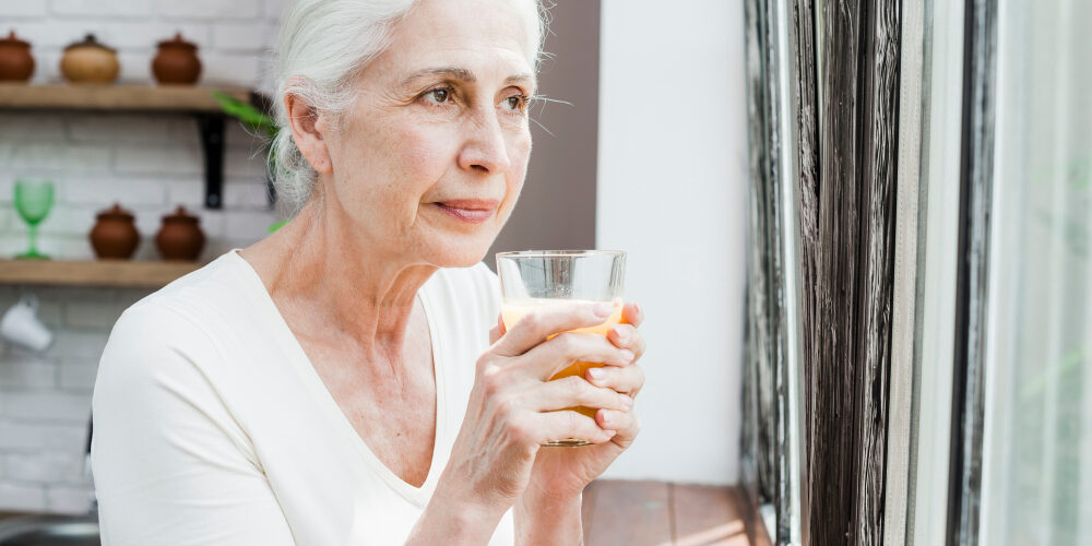 Elderly woman standing near the window of the house and holding glass with juice