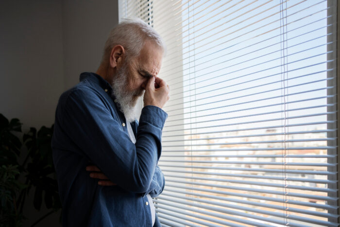 low vision difficulties Ηλικιωμένος άνδρας στέκεται κοντά στο παράθυρο, με σκυμμένο κεφάλι και ακουμπάει το χέρι στα μάτια του